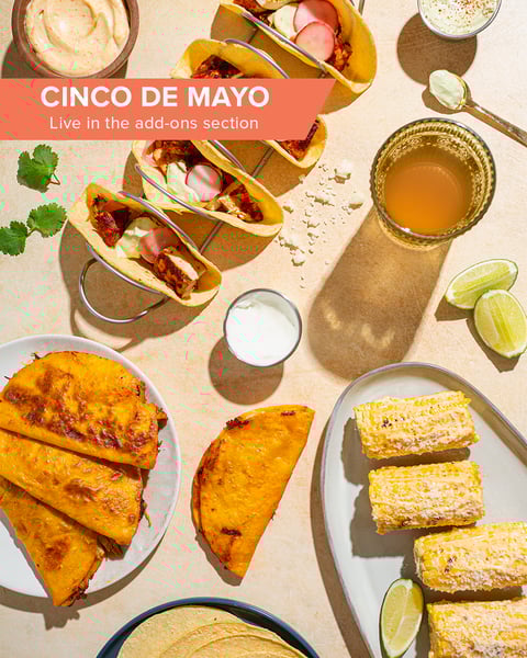A spread of tacos, elote, and a margarita, amidst lime wedges, cilantro, and crema dips. A text banner reads: Cinco de Mayo live in the add-ons section.